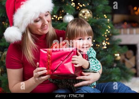 Glückliche Frau verleiht eingewickelt Weihnachten Geschenke Geschenke Kind Baby Kleinkind sitzen in der Nähe von Christmas tree Stockfoto