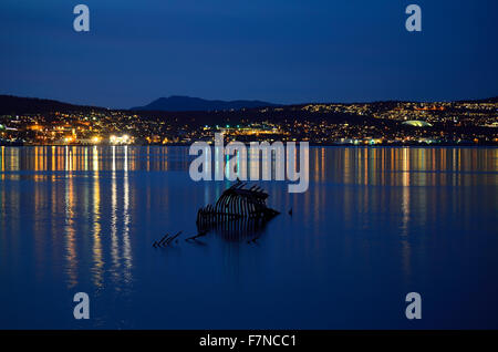 einsamer Kormoran Vogel sitzt auf einem verwitterten alten Holzschiff Wrack in cremigen weißen Fjords Wasser in der Nacht mit der Stadt-Insel tr Stockfoto