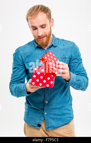 Aufgeregt, attraktive blonde jungen Mann mit Bart in Jeans Shirt Geschenk isoliert auf weißem Hintergrund öffnen Stockfoto