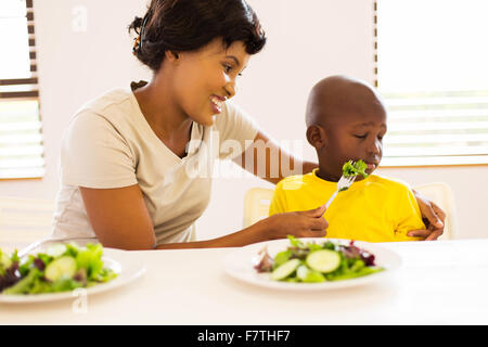 ziemlich afrikanischen Mutter versucht, ihren kleinen Sohn zu ernähren, der hasst, grünen Salat essen Stockfoto