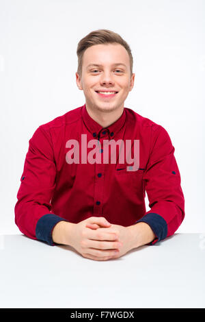Positiven fröhlicher junger Mann im roten Hemd sitzt und Kamera mit Blick auf weißem Hintergrund Stockfoto