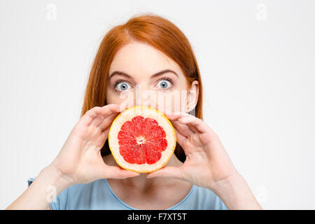 Porträt von schockiert amüsante junge Dame mit rotem Haar hält die Hälfte der Grapefruit vor den Mund über weißem Hintergrund Stockfoto