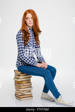 Nachdenkliche schöne lockige junge Frau mit langen roten Haaren im karierten Hemd und Jeans sitzen auf Bücher und denken über weiß Stockfoto