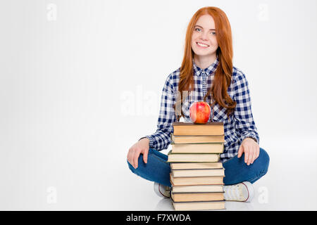 Positive, fröhliche junge rothaarige Frau sitzt mit den Beinen in der Nähe von Stapel Bücher mit Apple auf der oberen weißen Hintergrund gekreuzt Stockfoto