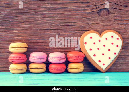 einige appetitlich Macarons mit verschiedenen Farben und Aromen und einem hausgemachten herzförmigen Cookie auf einer blauen Holzoberfläche gegen Stockfoto