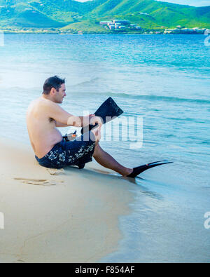 Ein Mann sitzt am Turtle Beach auf Buck Island National Monument mit St. Croix im Hintergrund und setzt auf Schwimmen flossen. Jungferninseln (US). USVI, U.S.V.I. Stockfoto