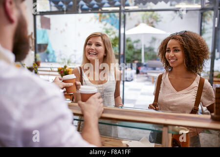 Indoor erschossen zwei junge Frauen Freunde Lächeln, wie sie ihre heiße Getränke aus dem Café-Zähler erhalten. Glückliche junge Freundinnen Stockfoto