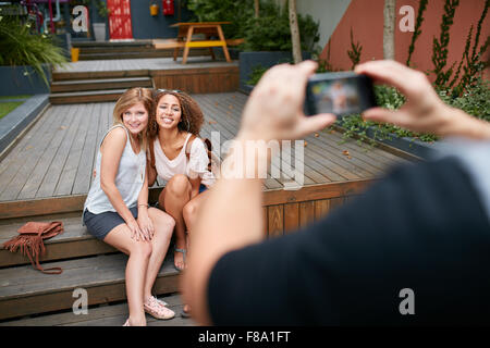 Zwei junge Frau sitzt zusammen posieren für ein Fotografieren ihres Freundes. Mann die Fotos seiner Freunde mit mob Stockfoto