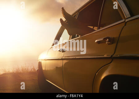 Schuss der Frau in einem Auto auf Reise entspannend. Weibliche Füße hängen aus Fahrzeugscheibe bei Sonnenuntergang. Stockfoto