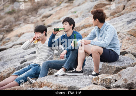 Drei junge Männer auf den Felsen sitzen und trinken Bier Stockfoto