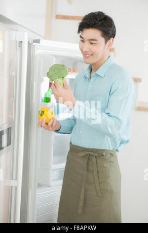 Lächelnd jungen Mann in Schürze mit Paprika und Brokkoli vor dem offenen Kühlschrank Stockfoto