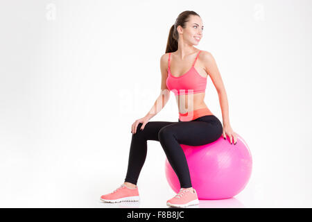 Fröhliche hübsche junge Fitness Frau sitzen auf rosa Fitball und Rückblick auf weißem Hintergrund Stockfoto