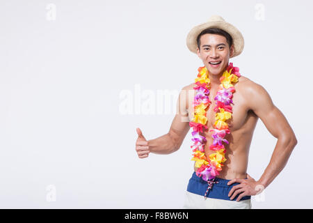 Muskulöser Mann im Schwimmen Hosen tragen Strohhut und Blume Halskette mit seinem Daumen oben und lächelnd Stockfoto