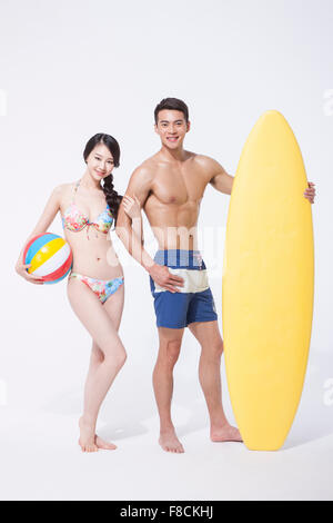 Paar in Strand tragen stehen zusammen halten, ein Beach-Ball und ein Surf-Brett jeweils beide starrte nach vorne mit einem Lächeln Stockfoto