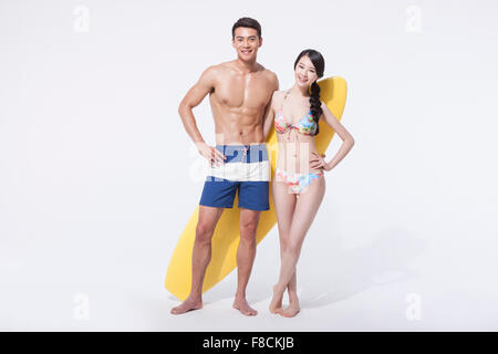 Paar in Strand zu tragen, zusammen mit einer Surf Board hinter ihnen mit ihrer Hand auf ihre Taille stehen und starrte nach vorne mit Stockfoto