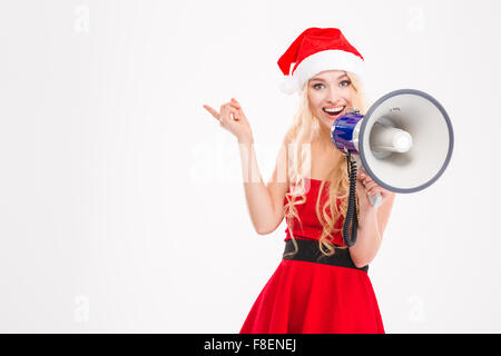 Schöne fröhliche junge Frau im Weihnachtsmann-Kostüm und Hut in Lautsprecher sprechen und über weißem Hintergrund Weg zeigend Stockfoto