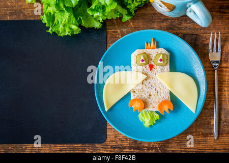 Gesundes Schulessen für Kinder. Lustige Eule Sandwich und Salat auf Holztisch plus leere schwarze Tafel für Text. Textfreiraum Stockfoto