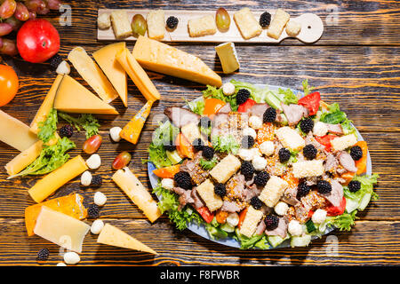 High Angle View of Gourmet-Salat mit frischem Gemüse, Brombeeren, Auswahl an Käse und Wurst mit Käse vom Brett und Zutaten auf rustikalen Holztisch. Stockfoto