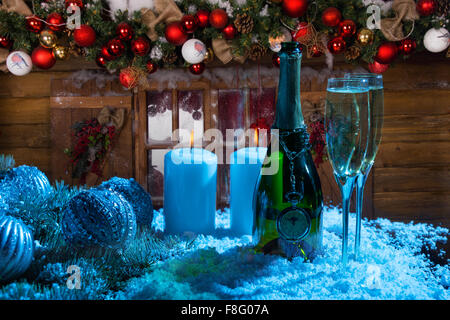 Festliche Weihnachten Stillleben in Blaulicht - Pocket Watch hängend Flasche von Champagner neben paar der Gläser auf verschneiten Oberfläche mit Kerzen beleuchtet, vor rustikale Holz-Hütte eingerichtet. Stockfoto