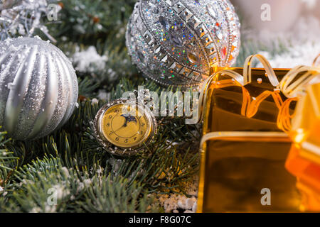 Festlichen Stillleben bedeckt von alten altmodischen Taschenuhr auf Schnee immergrüne Zweige mit funkelnden silberne Weihnachtskugeln mit Ecke des Geschenk verpackt in Goldpapier mit Schleife verziert. Stockfoto