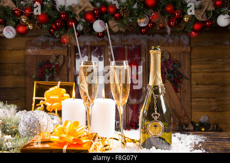 Festlichen Stillleben - Flasche Champagner mit zwei vollen Gläsern auf Schnee bedeckt Oberfläche mit Kerzen beleuchtet und Gold verpackt Geschenke vor rustikale Holz Kabine dekoriert zu Weihnachten. Stockfoto