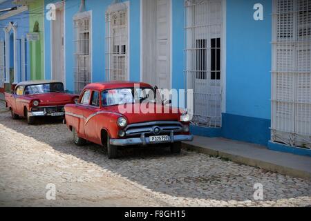 zwei rote Oldtimer abgestellt im Schatten auf einer gepflasterten Straße mit pastellfarbenen Gebäuden in Trinidad Kuba Stockfoto