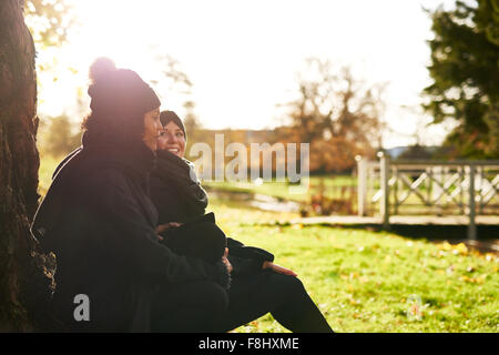 Zwei junge Frauen sitzen im herbstlichen Park, stützte sich auf Baumstamm. Sonnigen Stockfoto