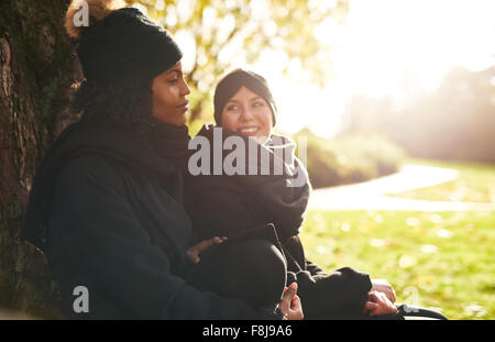 Zwei lächelnde junge Frauen sitzen im Park und am Baumstamm gelehnt. Sonnigen Stockfoto