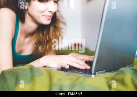 Junge Frau liegend auf Vorderseite auf Bett mit Laptop Computer lächelnd, differenzielle Fokus Stockfoto