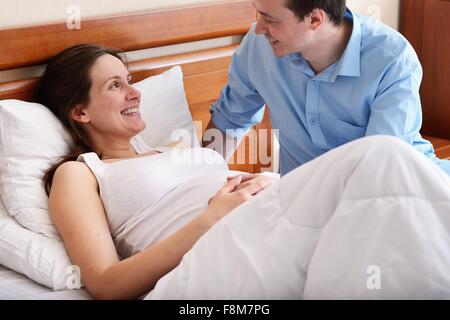 Schwangere Frau im Bett liegend, Mann neben ihr sitzen Stockfoto