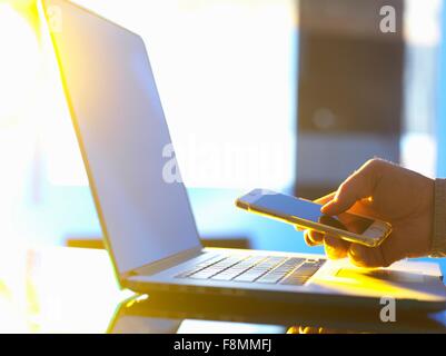 Geschäftsmann am Laptop-Computer mit Smartphone im Büro mit Sonnenlicht durch Fenster Stockfoto