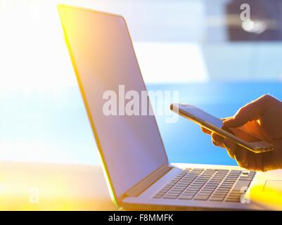Geschäftsmann am Laptop-Computer mit Smartphone im Büro mit Sonnenlicht durch Fenster Stockfoto