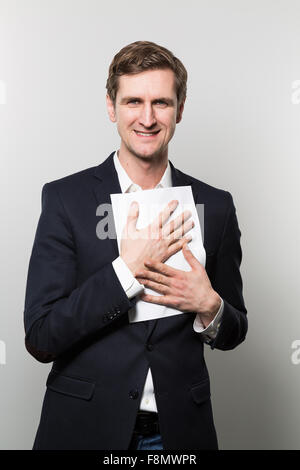 Blonden Geschäftsmann drückt ein Stück Papier an seine Brust mit einem frechen Grinsen während vor einem Farbverlauf Hintergrund Stockfoto
