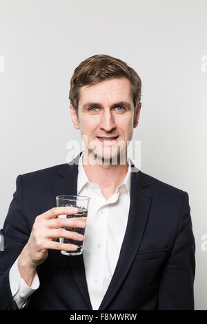 Blonden europäischen Geschäftsmann hält ein Glas Mineralwasser mit einem zufriedenen Blick vor einem Farbverlauf Hintergrund Stockfoto