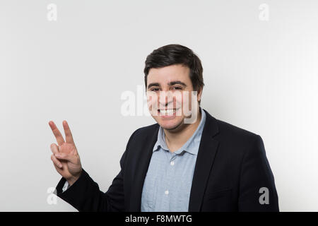 Dunkelhaarige Europäische Geschäftsmann mit einem breiten Grinsen macht ein Friedenszeichen während vor einem Farbverlauf Hintergrund Stockfoto