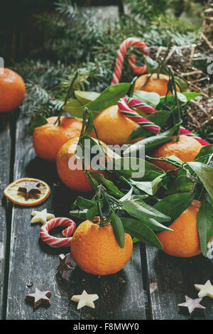 Mandarinen mit Blättern in Weihnachts-Dekor mit Weihnachtsbaum, trocken Orange und Bonbons über alten Holztisch. Stockfoto