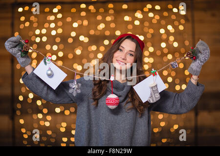 Attraktive positive junge Frau in Strickpullover und Fäustlinge mit handgemachten Weihnachtsschmuck über leuchtenden Hintergrund Stockfoto