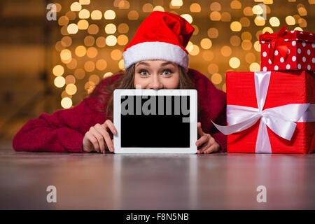 Amüsante positive Mädchen in Santa Claus Hut liegen und versteckt sich hinter leeren Tablettbildschirm über Urlaub Lichter Hintergrund Stockfoto