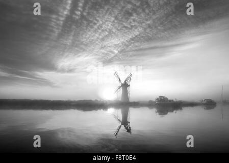 Atemberaubende Landschaft mit Windmühle und Fluss bei Sonnenaufgang am Sommermorgen in schwarz / weiß Stockfoto