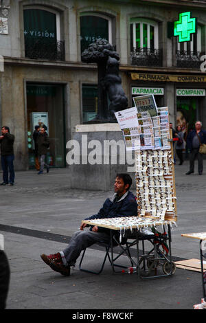 Madrid, Spanien 11. Dezember 2015: Ein Mann sitzt neben seinem Verkaufsstand und verkauft Tickets für die Weihnachtslotterie auf der Plaza Puerta del Sol im Zentrum von Madrid. Im Hintergrund sind der berühmte Bär und die Erdbeerbaum / Oso y el Madroño Statue, die ein Symbol von Madrid ist. Die spanische Weihnachtslotterie ist eine der ältesten der Welt und die größte der Welt in Bezug auf die Gesamtauszahlung. Das Siegerticket ist bekannt als „El Gordo“ („der große“ oder „der fette“). Quelle: James Brunker / Alamy Live News Stockfoto