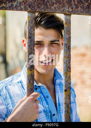 Hübsch lächelnd schwarz behaart, blau Augen jungen Mann im Hemd hinter Käfig oder Tor Metallstangen Stockfoto