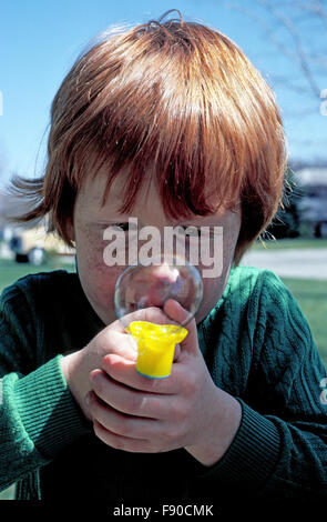 Ein netter vier-jährige amerikanische junge mit roten Haaren und einem sommersprossiges Gesicht bläst eine Seifenblase aus dem gelben Rohr beim Spielen im Freien in seinen grünen Pullover.  -Modell veröffentlicht. Stockfoto