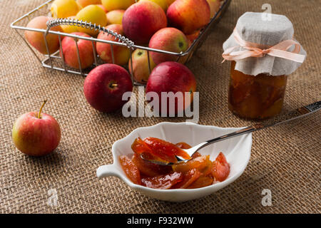 Hausgemachte Apfel Marmelade auf weiße Untertasse, Löffel, Glas Marmelade und Äpfel in Metall Drahtkorb auf einem Tisch mit Sackleinen abgedeckt. Sele Stockfoto