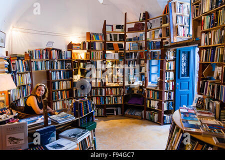 Santorini, Oia. Atlantis second hand Buchladen. Interieur, Wände in Holz Regale voller Bücher, Frau shop Assistent am Schreibtisch sitzen, ein Lächeln auf den Lippen. Stockfoto