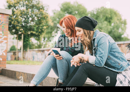 Zwei junge hübsche kaukasischen Blonde und Rothaarige glattes Haar Frauen sitzen auf einer Treppe mit Smartphone, auf der Suche nach unten den Bildschirm - soziales Netzwerk, Technik, Kommunikations-Konzept - Seitenansicht Stockfoto
