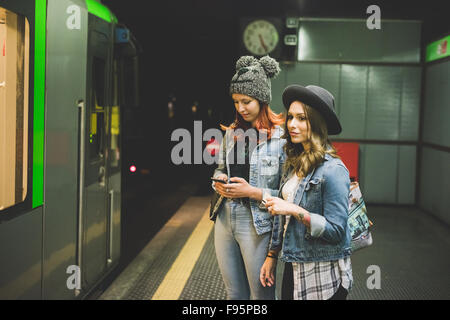 Knie Abbildung zweier junger hübscher kaukasischen Blonde und Rothaarige glattes Haar Frauen warten im Untergrund für Rohr, mit Smartphone - Transport, Technologie-Konzept Stockfoto
