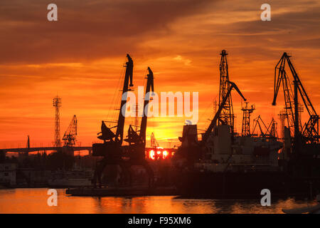 Silhouetten von Kränen und Frachtschiffe im Hafen von Varna bei Sonnenuntergang unter leuchtend roter Himmel Stockfoto