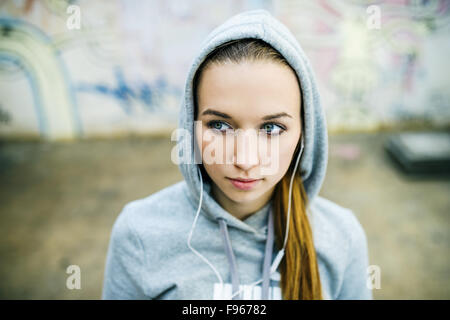 Schwere Teenager-Mädchen mit Kapuze auf anhören von Musik über Kopfhörer auf Hintergrund der Graffitiwand stehend Stockfoto