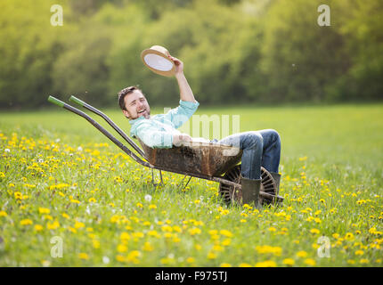 Junger männliche Bauer ist entspannend in Schubkarre auf der grünen Wiese Stockfoto