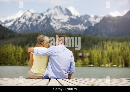 Senior paar sitzt am Pier über dem Bergsee mit den Bergen im Hintergrund Stockfoto